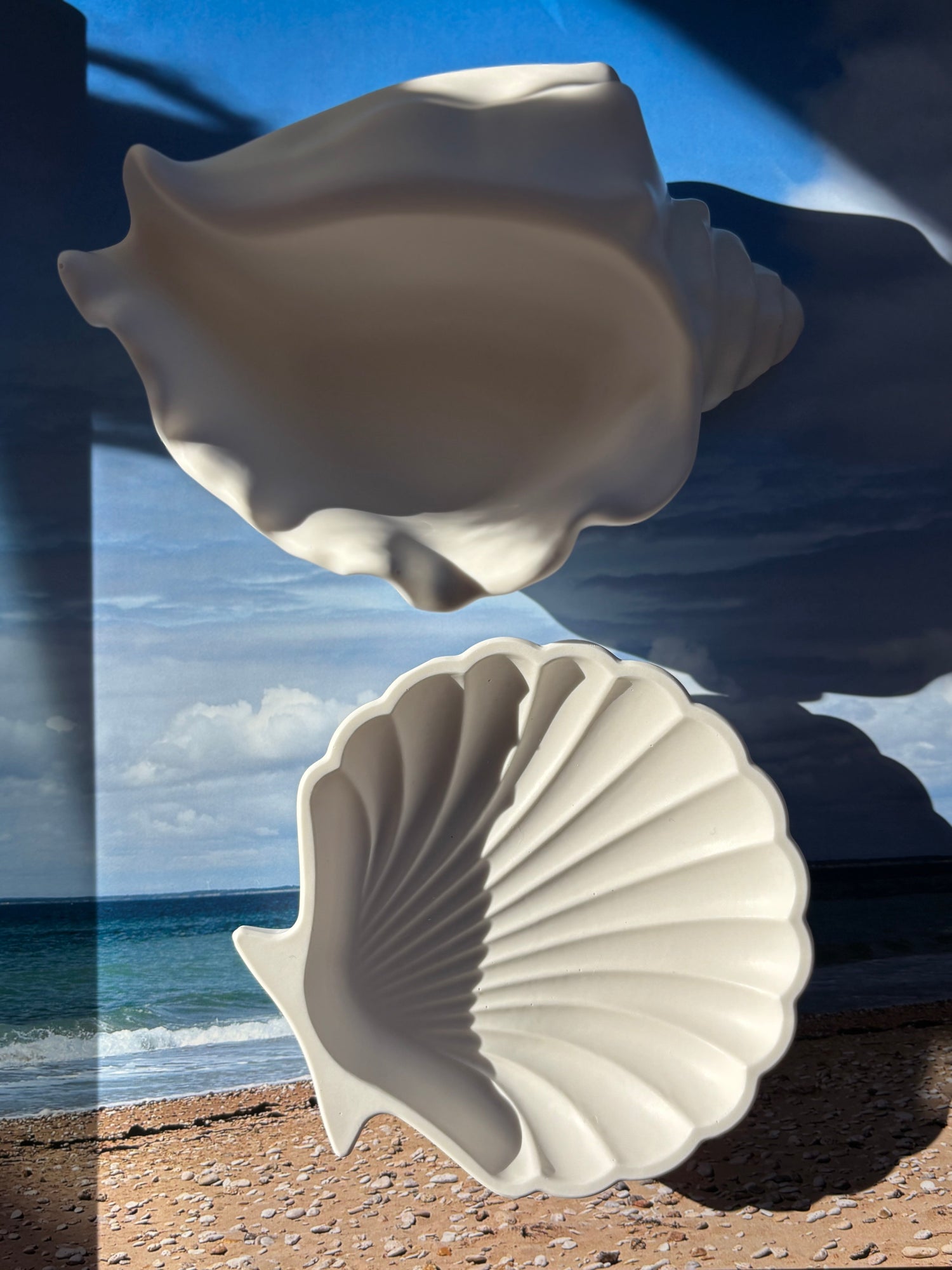 Two sculptural white shell-shaped trays displayed against a serene beach backdrop, evoking summer and tranquility.